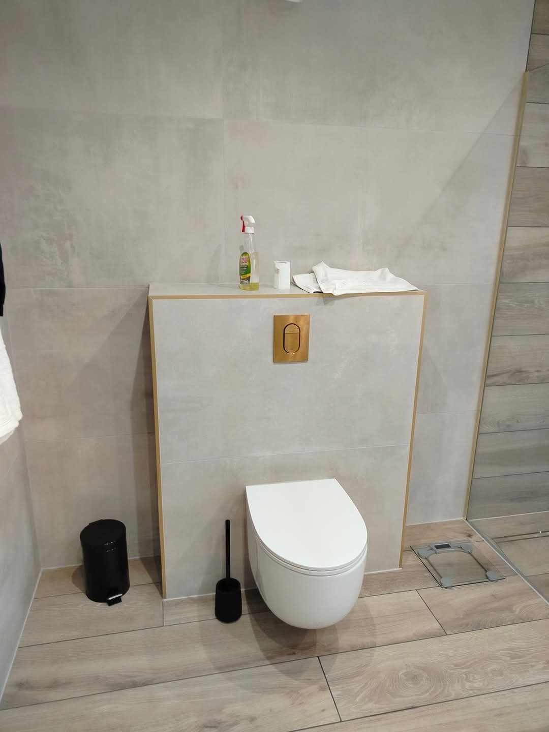 Nowoczesna toaleta podwieszana z zabudową w kolorze jasnego betonu, złotym przyciskiem spłukiwania i drewnopodobną podłogą; widoczne akcesoria: czarny kosz na śmieci i szczotka do WC, oraz środki...