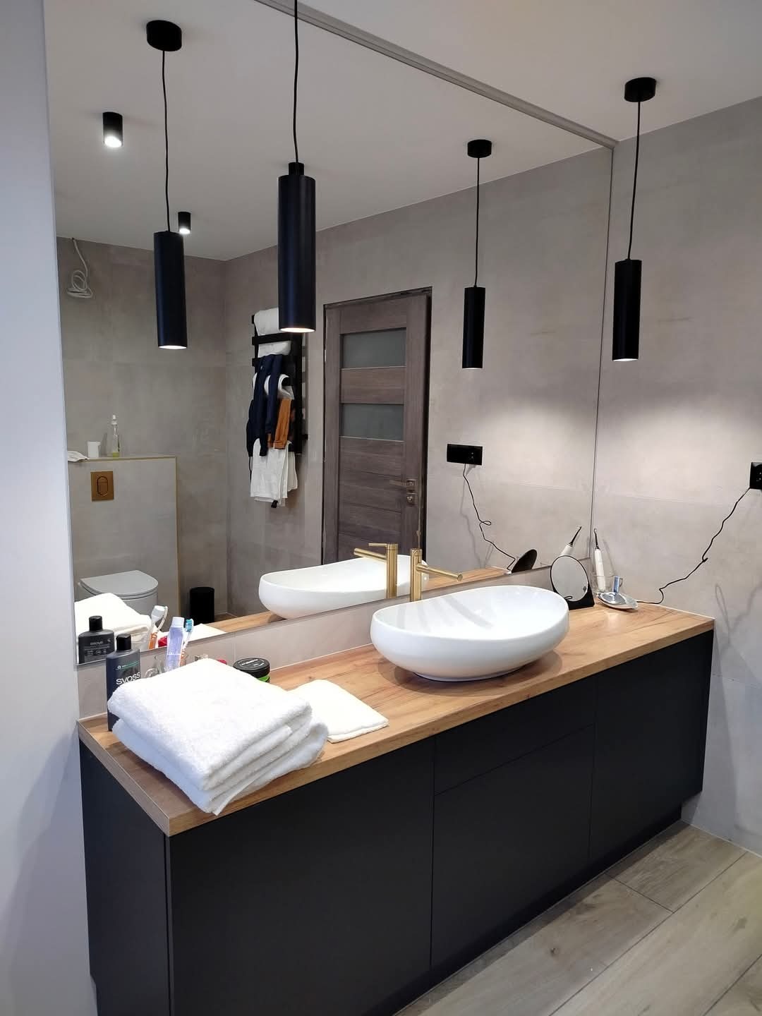 Nowoczesna łazienka z owalną umywalką nablatową, złotą baterią, czarnymi lampami wiszącymi i dużym lustrem na ścianie z szarymi płytkami.