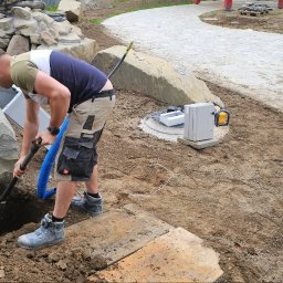 MARCIN BOŁOZ - Pracownik kopie dół obok dużego kamienia, przygotowując miejsce na instalację elektryczną w ogrodzie. Widoczna skrzynka elektryczna i narzędzia.