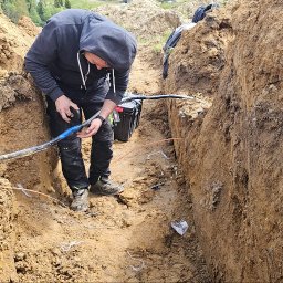 MARCIN BOŁOZ - Elektryk łączy kabel w wykopie ziemnym, używając taśmy izolacyjnej. Widoczna skrzynka narzędziowa, fragment krajobrazu i ściany wykopu.
