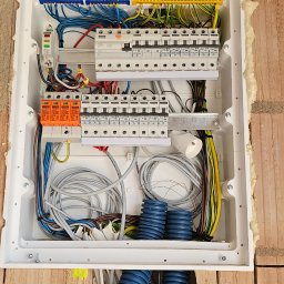 MARCIN BOŁOZ - Rozdzielnica elektryczna wmurowana w ścianę z cegły, zabezpieczona pianką montażową, z widocznymi przewodami i niebieskimi rurkami karbowanymi.