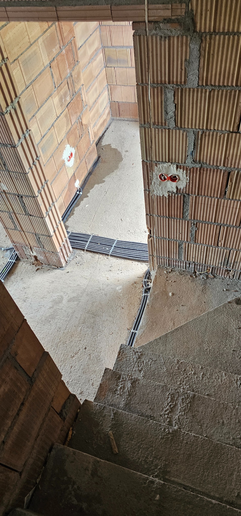 Wnętrze w trakcie budowy: ceglane ściany, instalacja elektryczna w peszlach na podłodze i w ścianach, surowy beton schodów. Widoczne puszki elektryczne w ścianie.