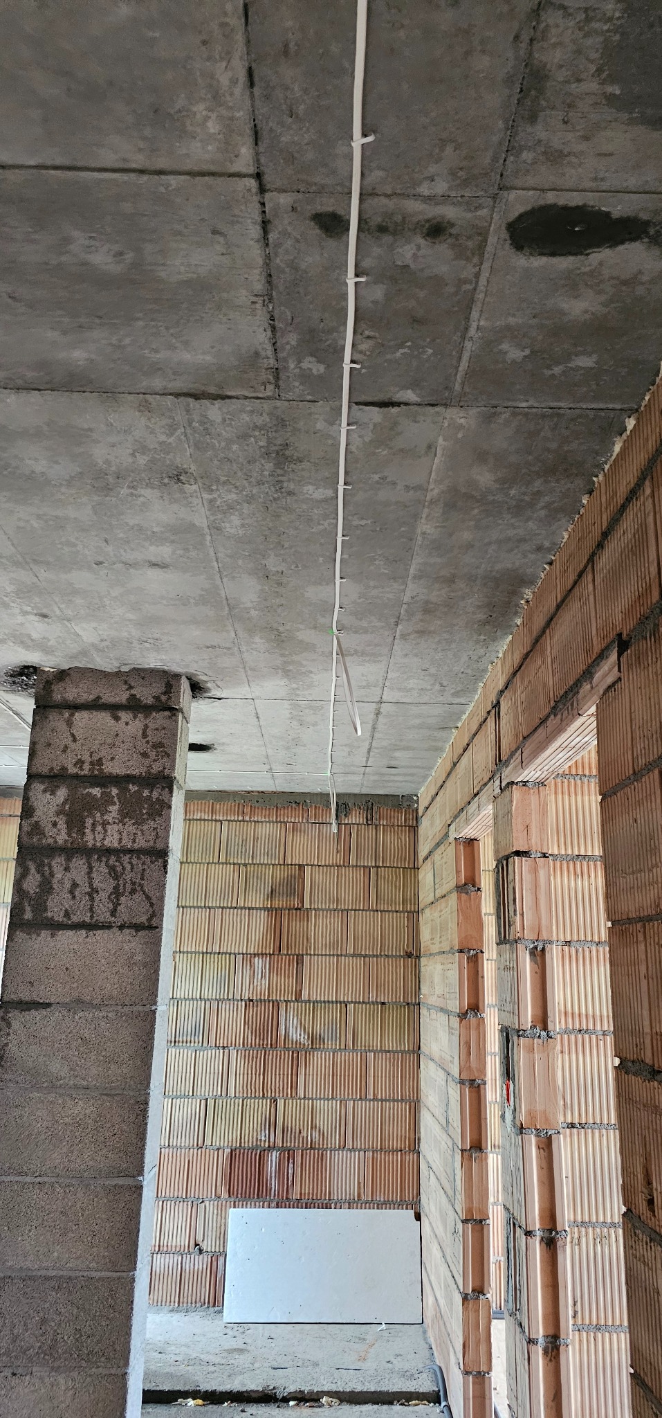 Surowy strop betonowy z przymocowanym kablem elektrycznym, ściany z czerwonej cegły dziurawki w stanie surowym i betonowy filar w nowym budynku.