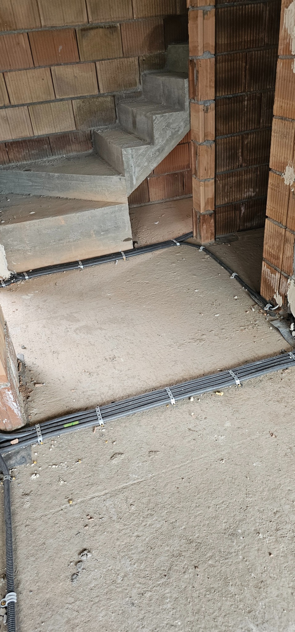 Instalacja elektryczna w peszlach, prowadzona po podłodze w surowym stanie budynku, przed wylaniem posadzki. Widoczne schody i ściany z cegły.