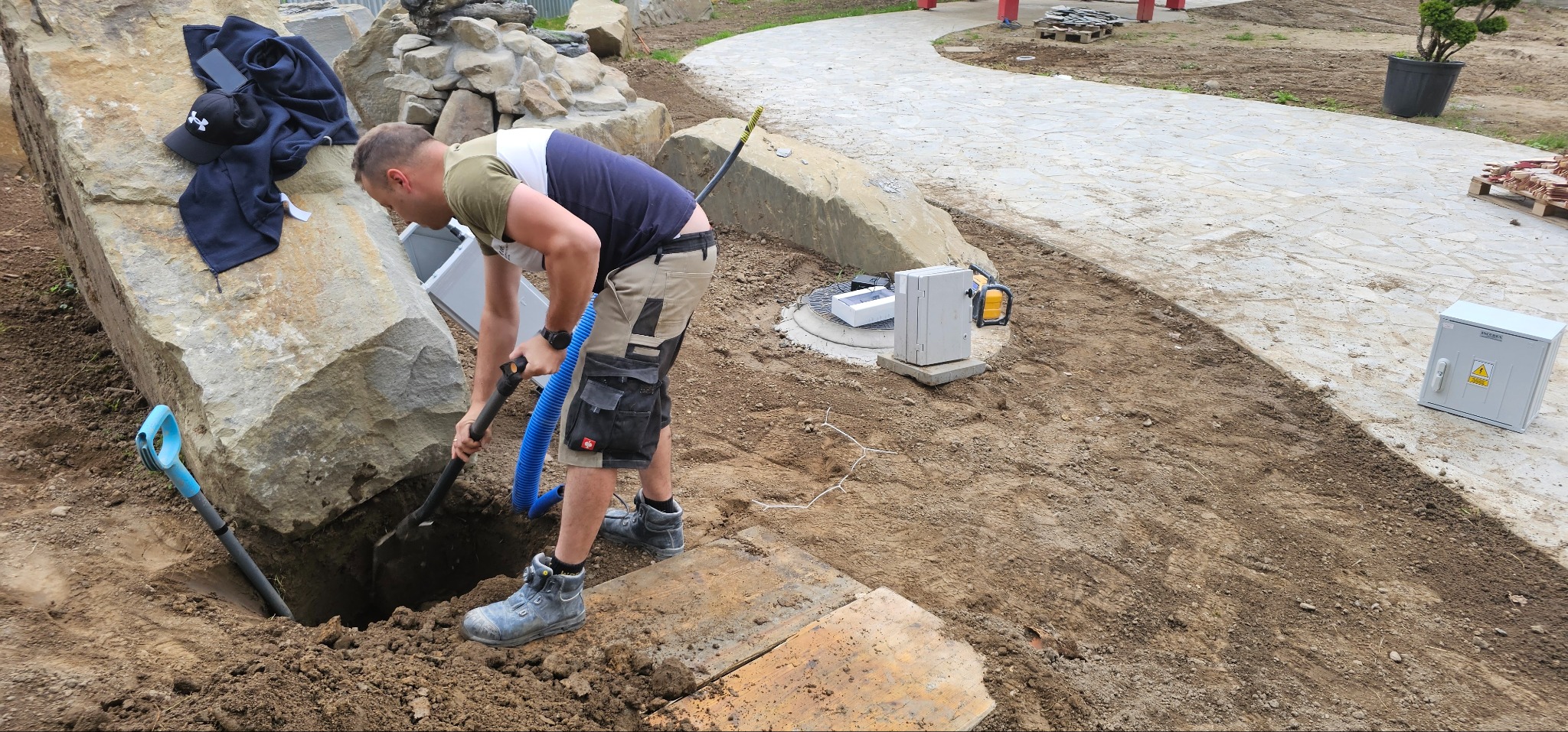 Pracownik kopie dół obok dużego kamienia, przygotowując miejsce na instalację elektryczną w ogrodzie. Widoczna skrzynka elektryczna i narzędzia.