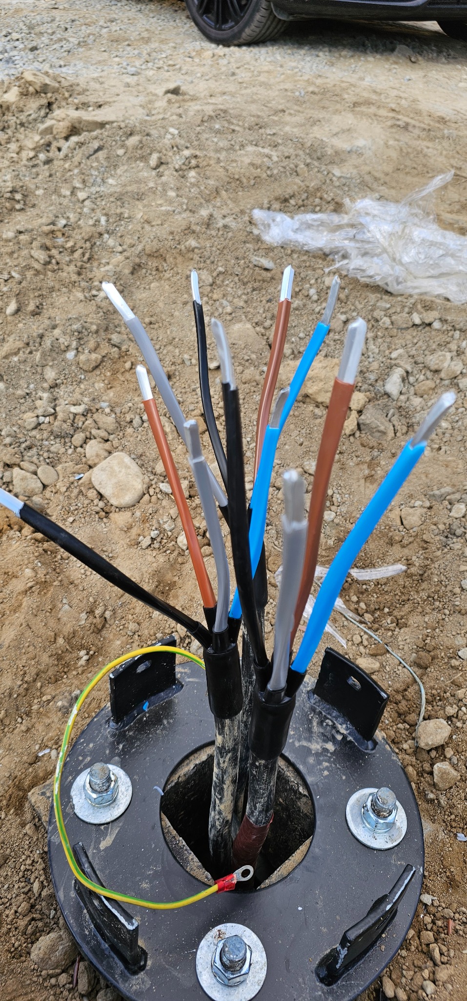 Przyłącze kablowe z widocznymi przewodami o różnych kolorach, wystającymi z czarnej, okrągłej obudowy. Widoczne ślady ziemi i kamieni.