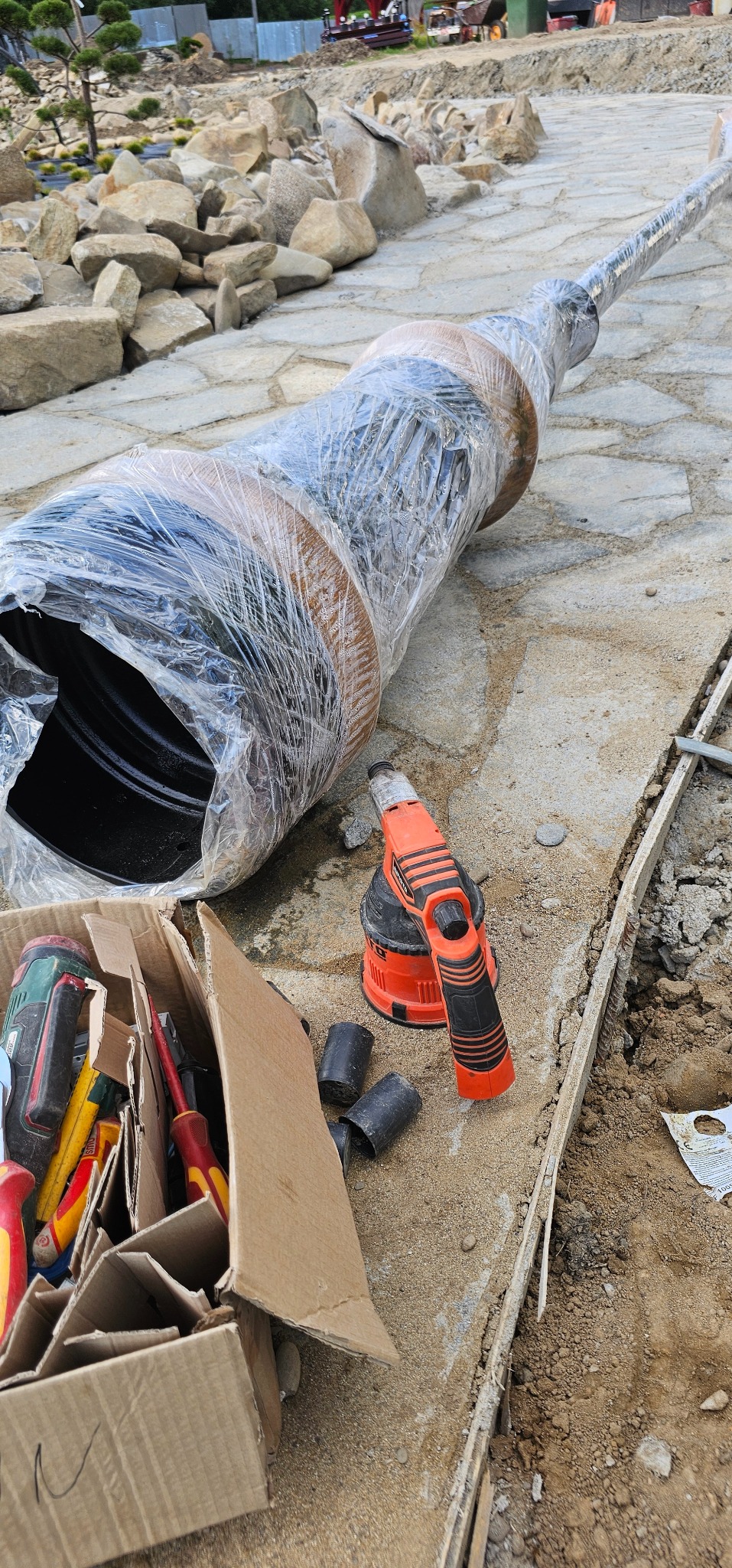 Długa rura w folii na placu budowy, obok narzędzia i karton ze śrubokrętami. Widoczne kamienie i elementy krajobrazu, sugerujące prace ziemne i instalacyjne.