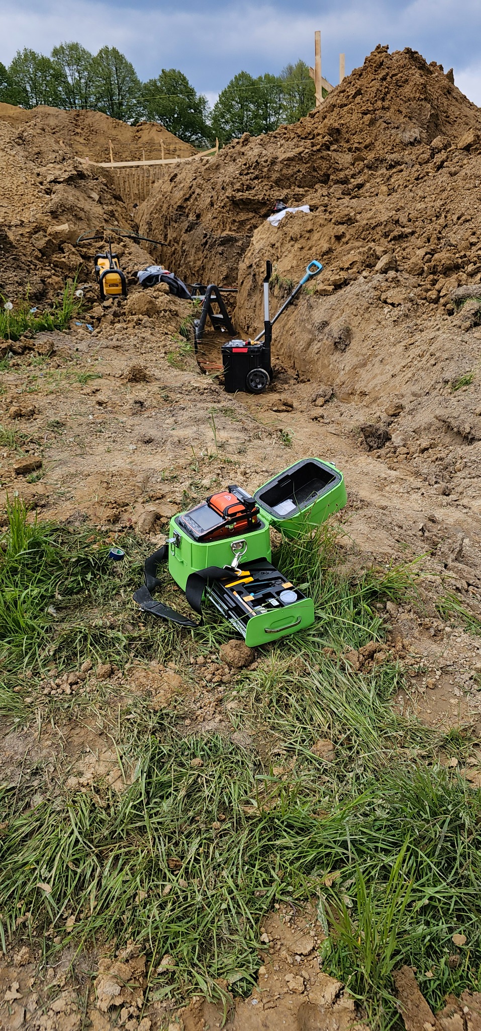 Otwarta skrzynka narzędziowa z narzędziami na trawie, w tle wykop ziemny z widocznymi elementami instalacji. Sprzęt pomiarowy w skrzynce.