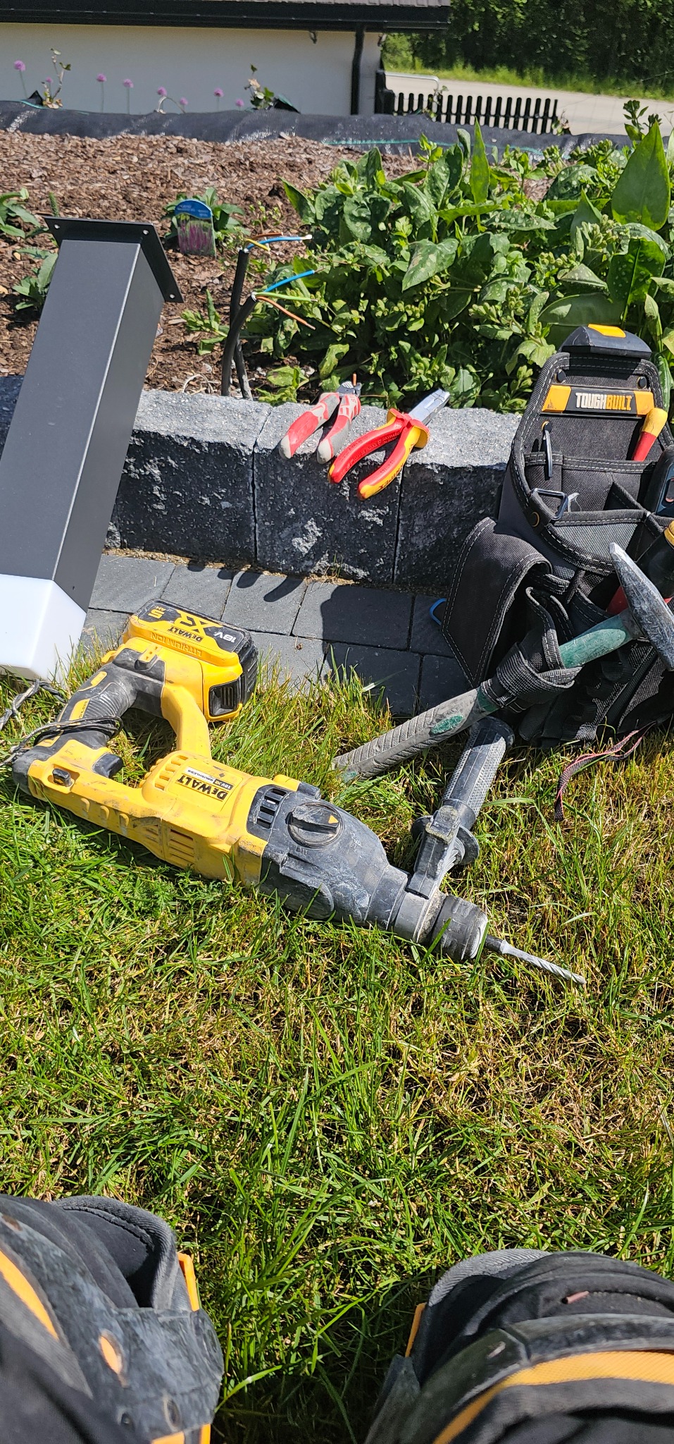 Wiertarka udarowa DeWalt na trawie obok narzędzi i słupka oświetleniowego. Widoczne rękawice robocze i torba narzędziowa Toughbuilt.