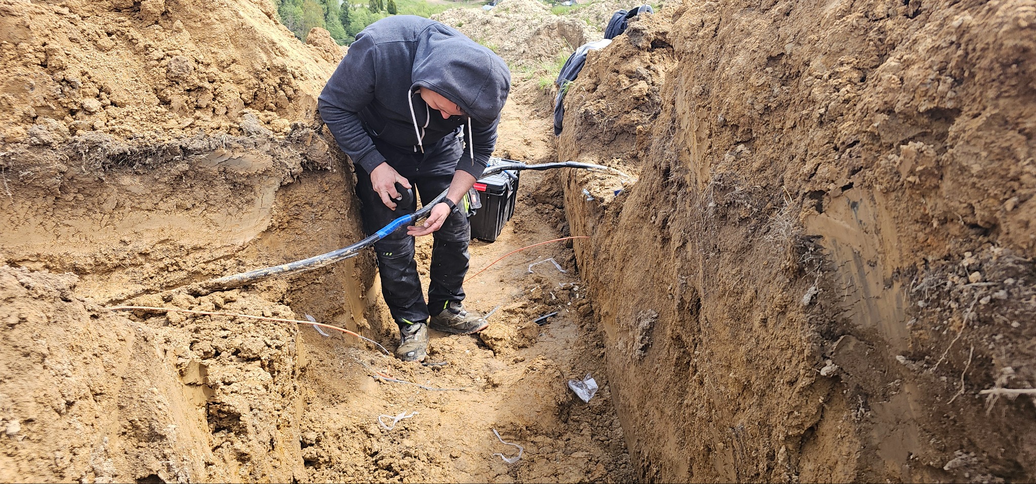 Elektryk łączy kabel w wykopie ziemnym, używając taśmy izolacyjnej. Widoczna skrzynka narzędziowa, fragment krajobrazu i ściany wykopu.