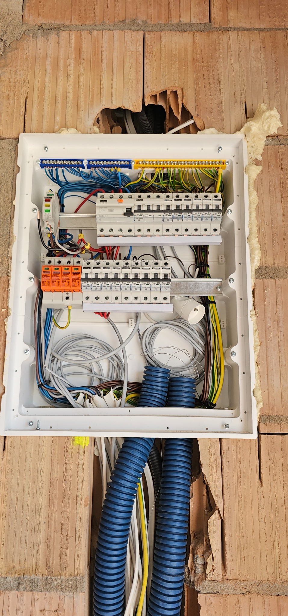 Rozdzielnica elektryczna wmurowana w ścianę z cegły, zabezpieczona pianką montażową, z widocznymi przewodami i niebieskimi rurkami karbowanymi.