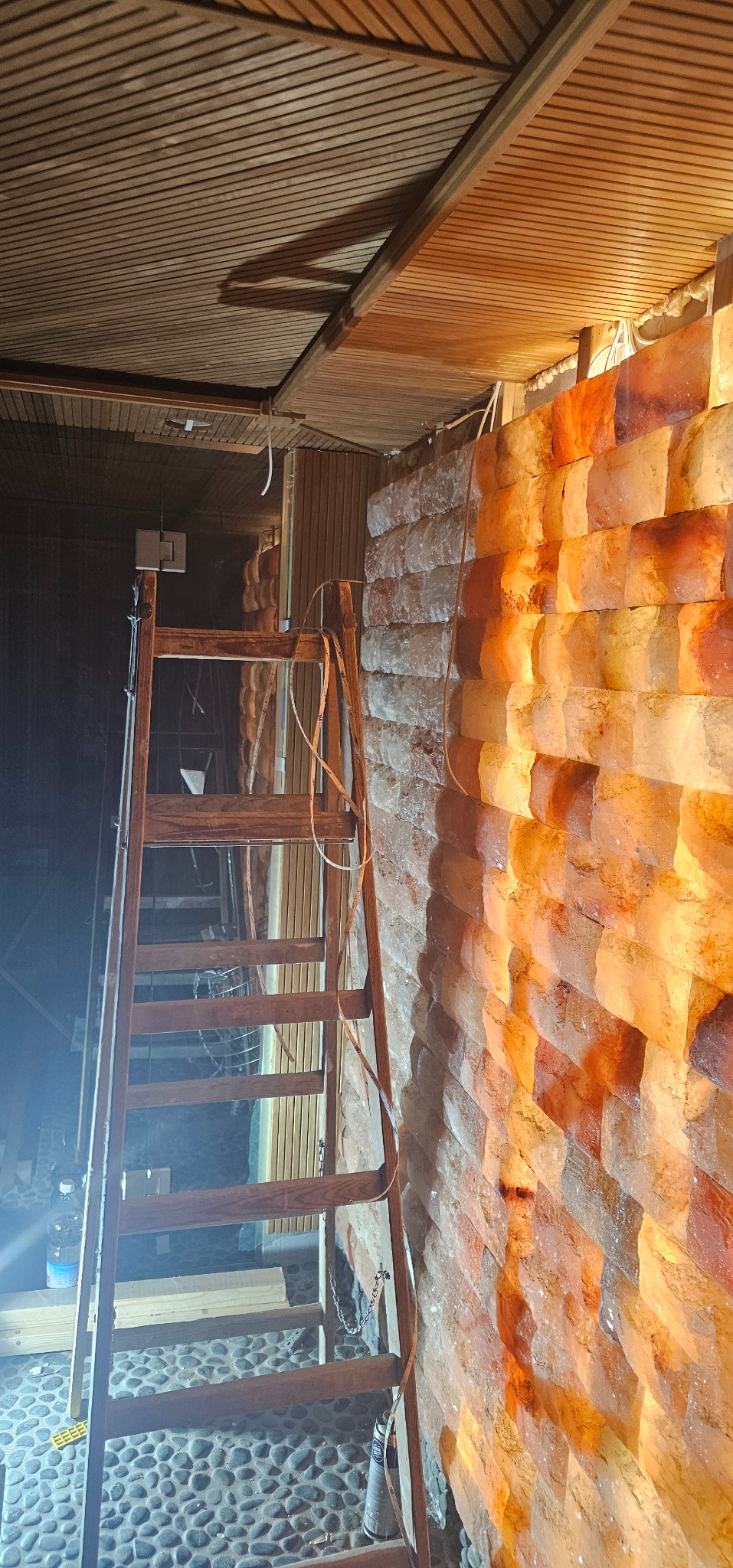 Montaż ściany z bloków solnych w saunie, widoczna drewniana drabina, elementy instalacji elektrycznej i drewniane wykończenie sufitu.