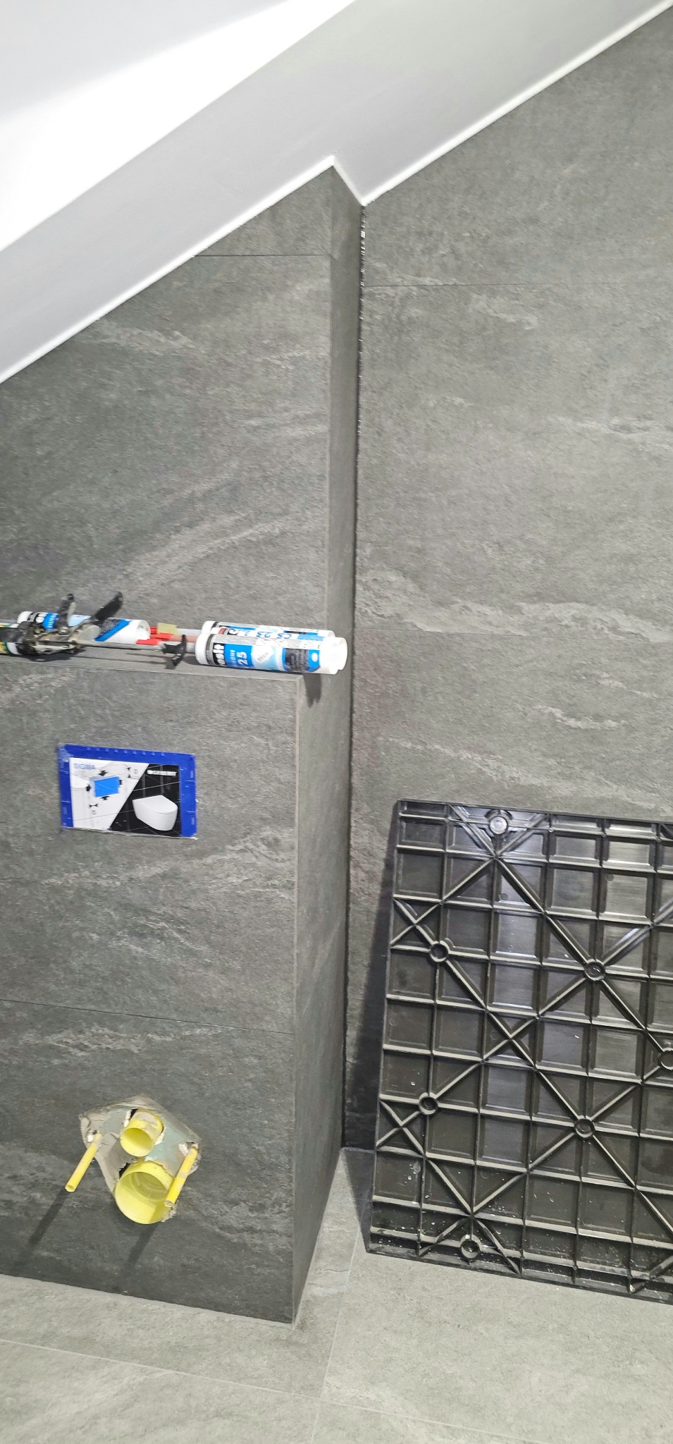 Szare płytki imitujące kamień na ścianie w łazience, widoczne narzędzia i elementy instalacji podtynkowej WC. Ukończone fugowanie narożników.