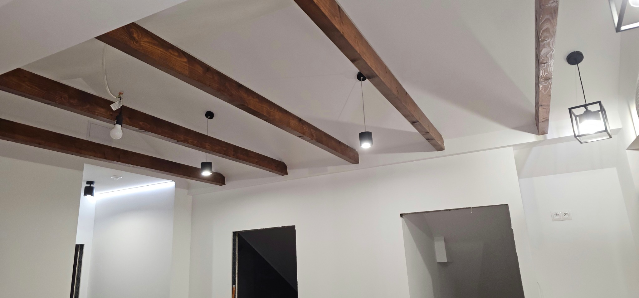 Białe wnętrze z drewnianymi belkami stropowymi i nowoczesnym oświetleniem. Widoczne gniazdka elektryczne i otwory w ścianach.