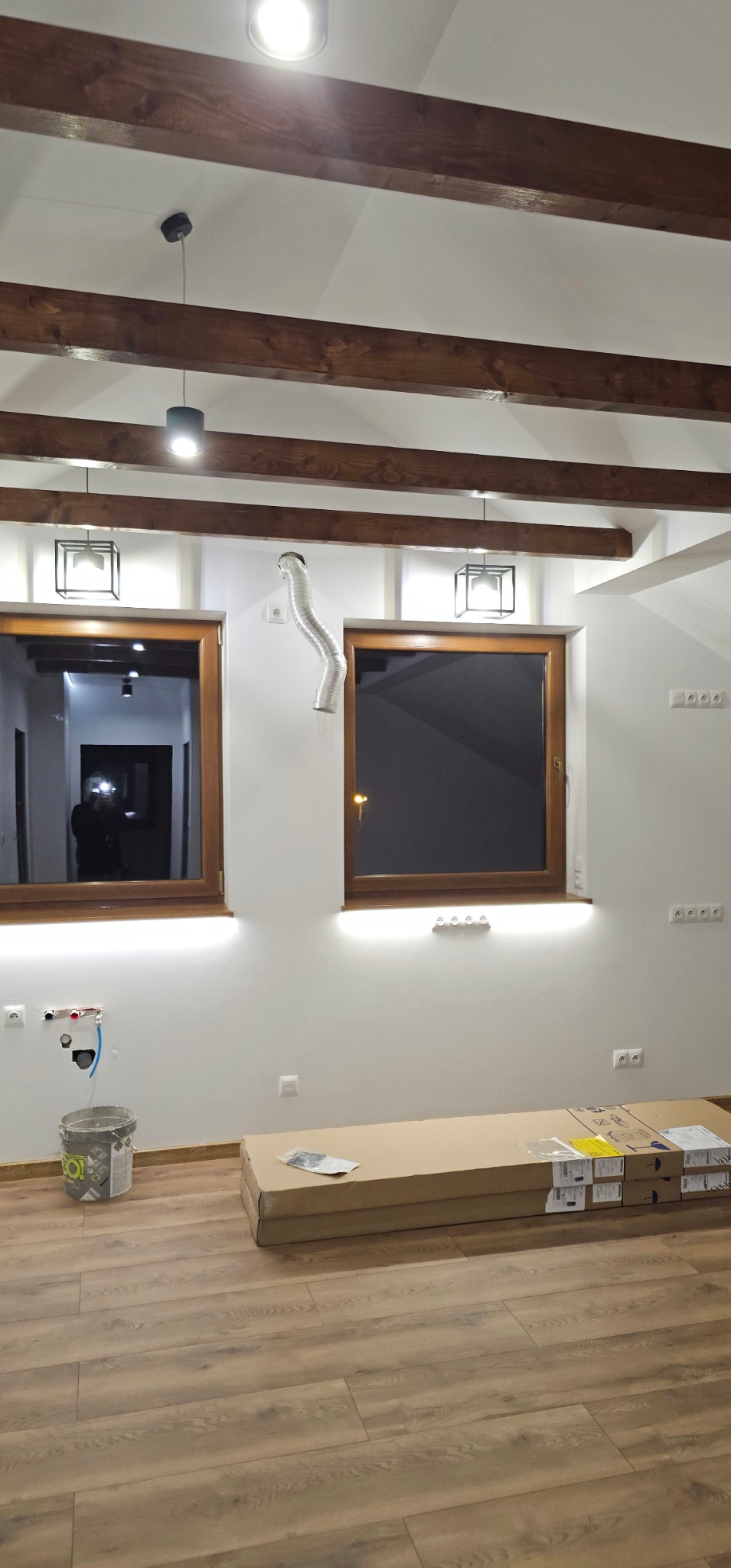 Wnętrze w trakcie wykańczania: drewniane belki stropowe, okna z podświetleniem LED, widoczne puszki elektryczne, rura wentylacyjna i kartony na podłodze.