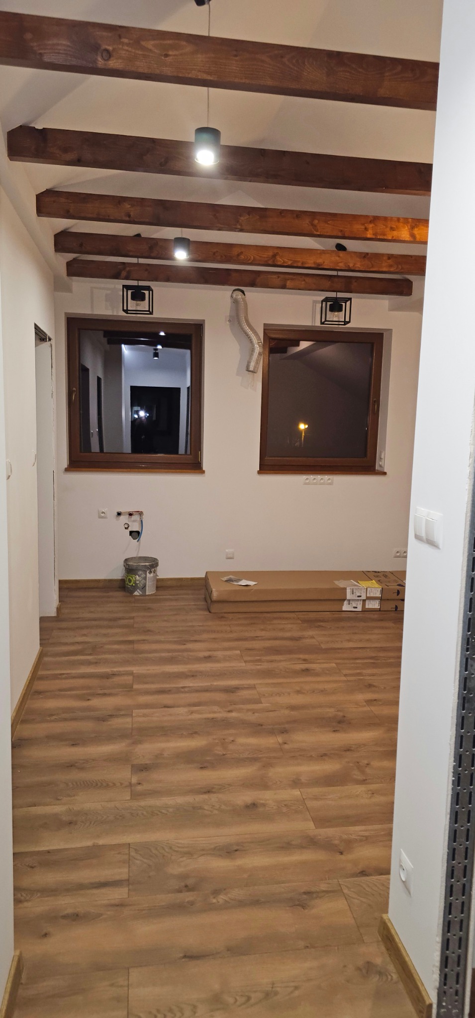 Wnętrze po remoncie z drewnianą podłogą, widoczne okna, drewniane belki na suficie i lampy. Na podłodze leżą panele w kartonach i stoi wiadro z farbą.