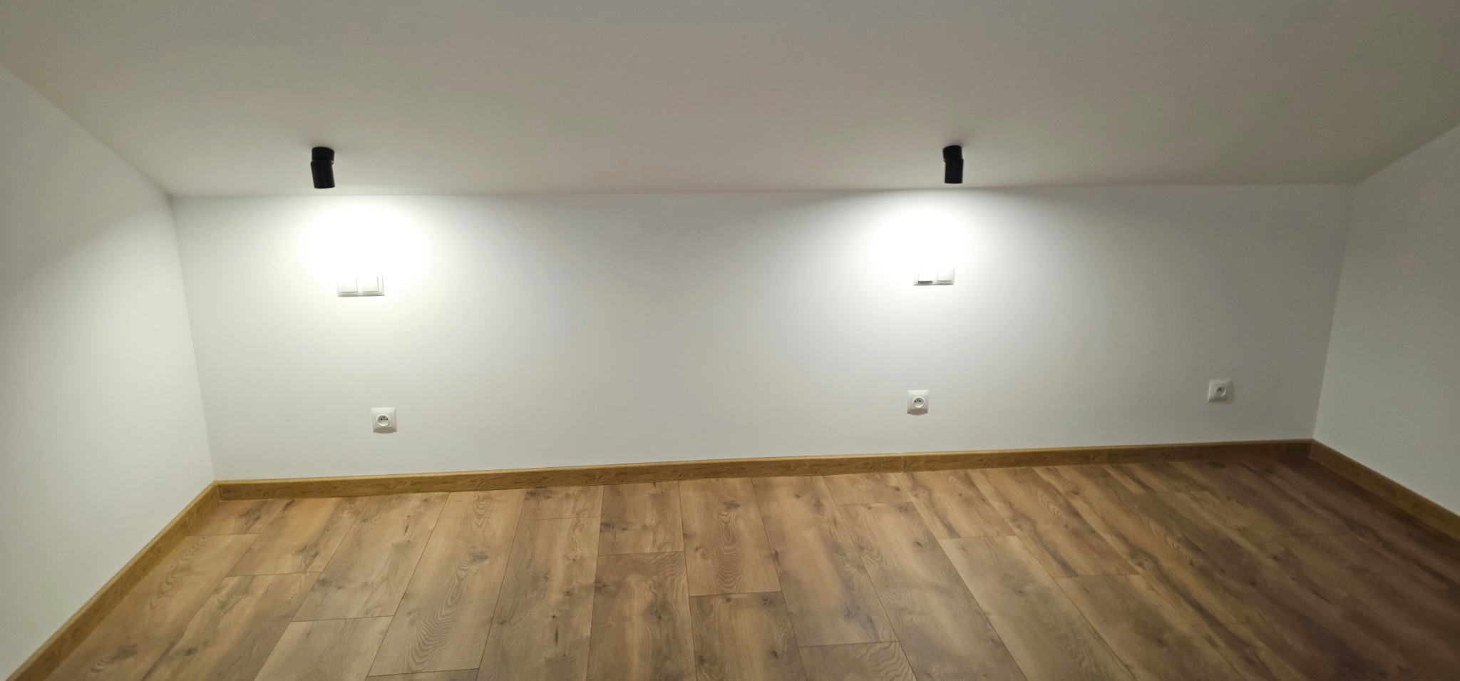 Jasne wnętrze po wykończeniu: drewnopodobna podłoga, białe ściany z gniazdkami i włącznikami, czarne lampy sufitowe. Minimalistyczny design, świeżo po remoncie.
