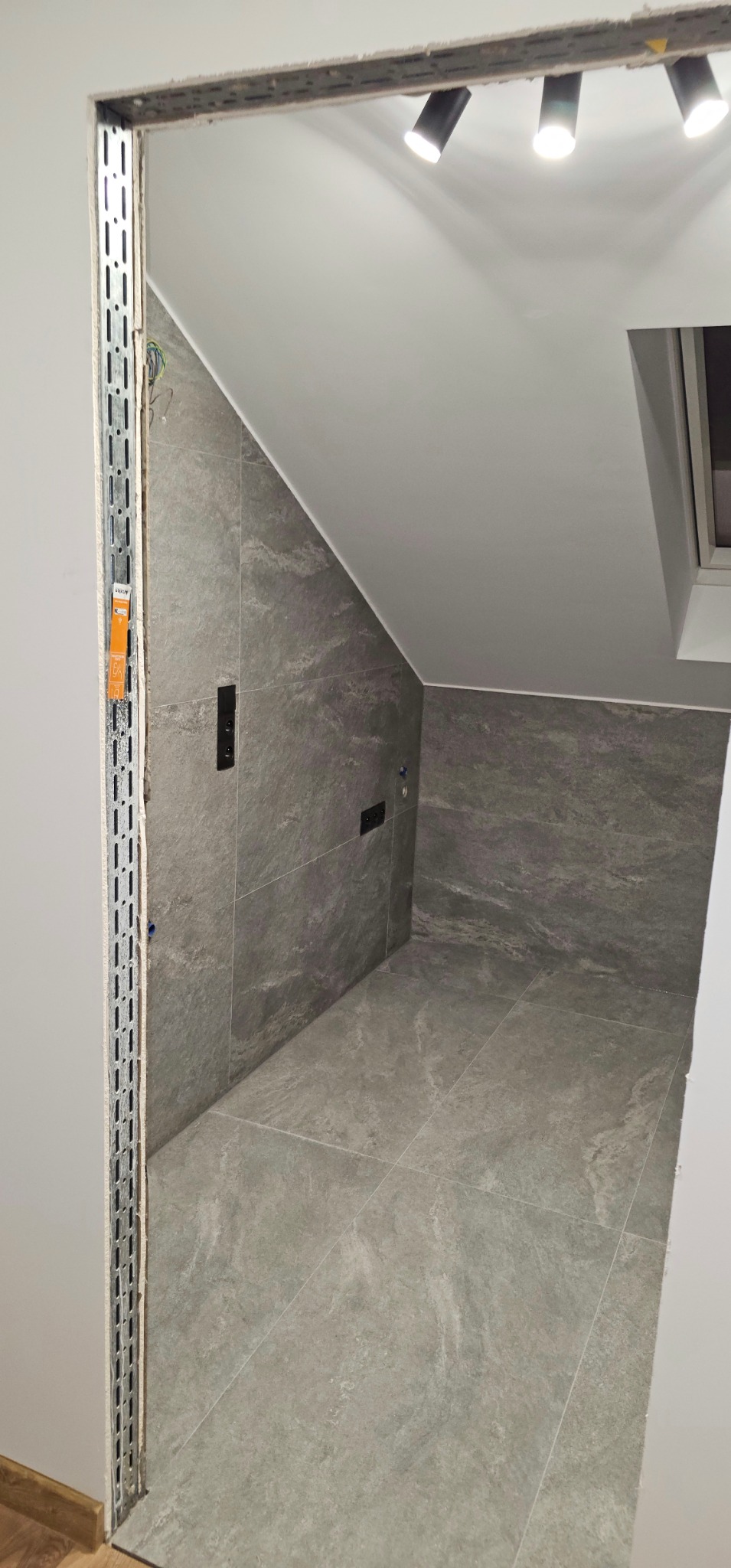 Wykończona łazienka w szarościach z oświetleniem punktowym i widoczną instalacją elektryczną w ścianie. Ukończone płytki na podłodze i ścianach.