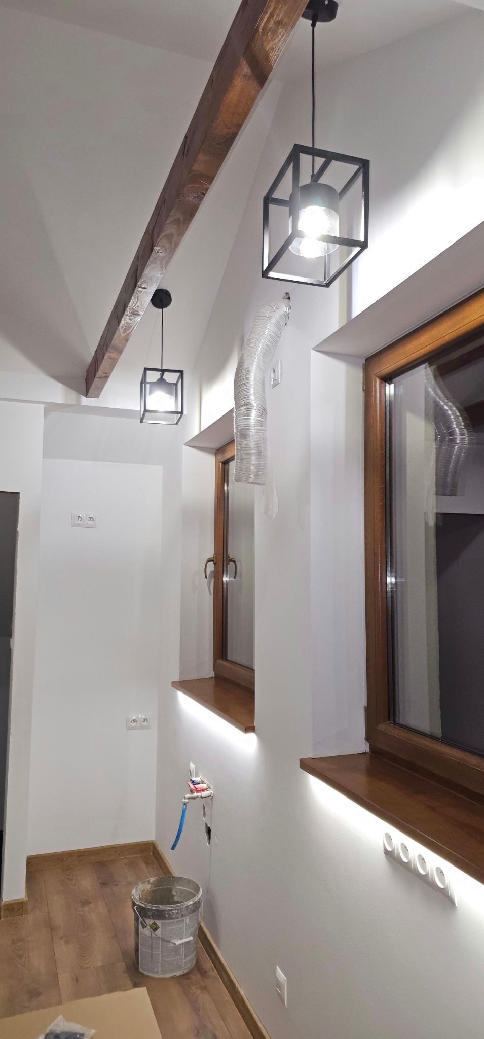 Wnętrze po remoncie: drewniana belka stropowa, nowoczesne lampy w kształcie sześcianu, okna z podświetlanymi parapetami LED, gładkie, białe ściany z gniazdkami.