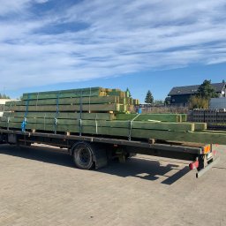 APHD D.M.K. KAWIŃSCY SPÓŁKA CYWILNA - Ciężarówka z załadunkiem impregnowanych belek drewnianych, zabezpieczonych niebieskimi pasami transportowymi, na tle błękitnego nieba z chmurami i zabudowań.