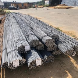 APHD D.M.K. KAWIŃSCY SPÓŁKA CYWILNA - Sztaple wiązek prętów zbrojeniowych z etykietami producenta, ułożone na placu budowy, w tle budynki i osoba w odblaskowej kamizelce.