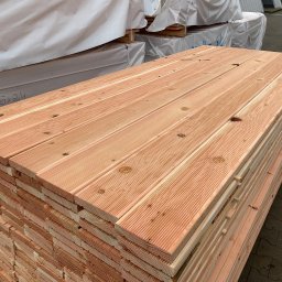 APHD D.M.K. KAWIŃSCY SPÓŁKA CYWILNA - Stos desek tarasowych z jasnego drewna, widoczne słoje i sęki, pakowane w białą folię ochronną, ułożone na zewnątrz.