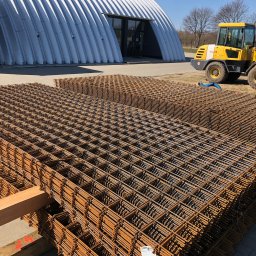 APHD D.M.K. KAWIŃSCY SPÓŁKA CYWILNA - Stosy siatek zbrojeniowych na drewnianych legarach, w tle hala magazynowa o łukowej konstrukcji i żółta ładowarka Komatsu WA75.