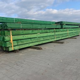 APHD D.M.K. KAWIŃSCY SPÓŁKA CYWILNA - Stos impregnowanych na zielono belek drewnianych, zabezpieczonych żółtą taśmą, składowanych na placu firmy w Drawsku Pomorskim, widok z boku, w tle błękitne niebo z chmurami.