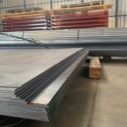 APHD D.M.K. KAWIŃSCY SPÓŁKA CYWILNA - Stos arkuszy blachy stalowej na drewnianej podkładce w magazynie, w tle ułożone palety i profile stalowe.