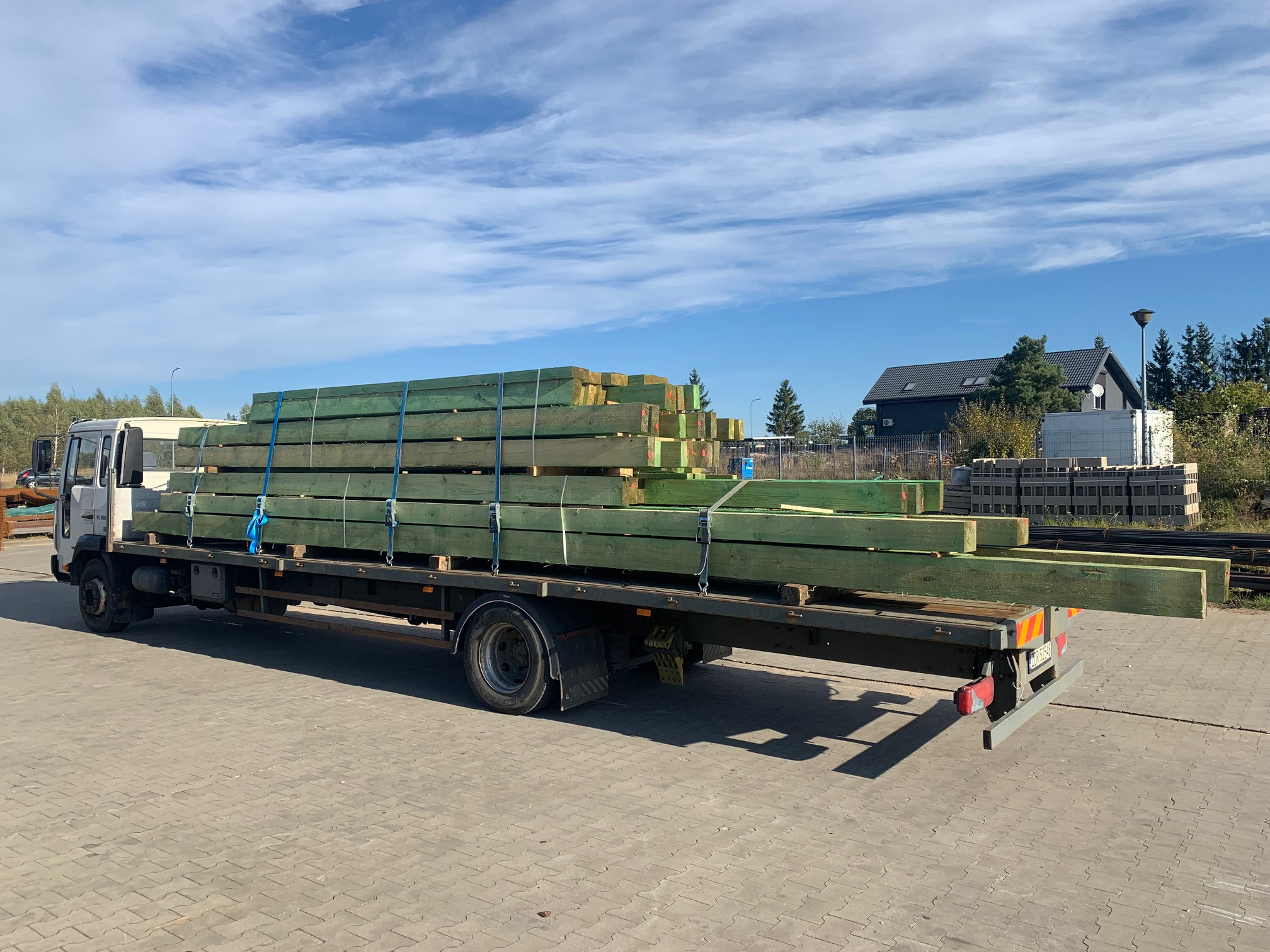 Ciężarówka z załadunkiem impregnowanych belek drewnianych, zabezpieczonych niebieskimi pasami transportowymi, na tle błękitnego nieba z chmurami i zabudowań.