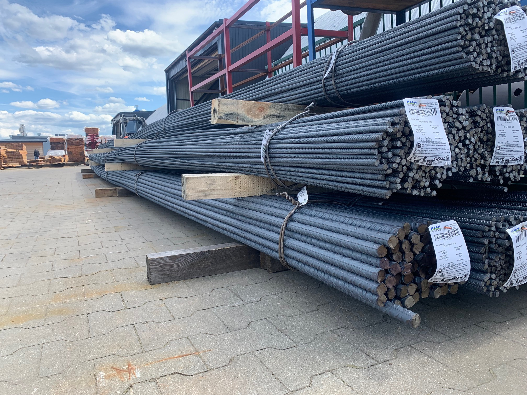 Stosy wiązek prętów zbrojeniowych na drewnianych legarach, z etykietami producenta, ułożone na placu budowy z kostki brukowej, z widocznym w tle składem drewna i błękitnym niebem.