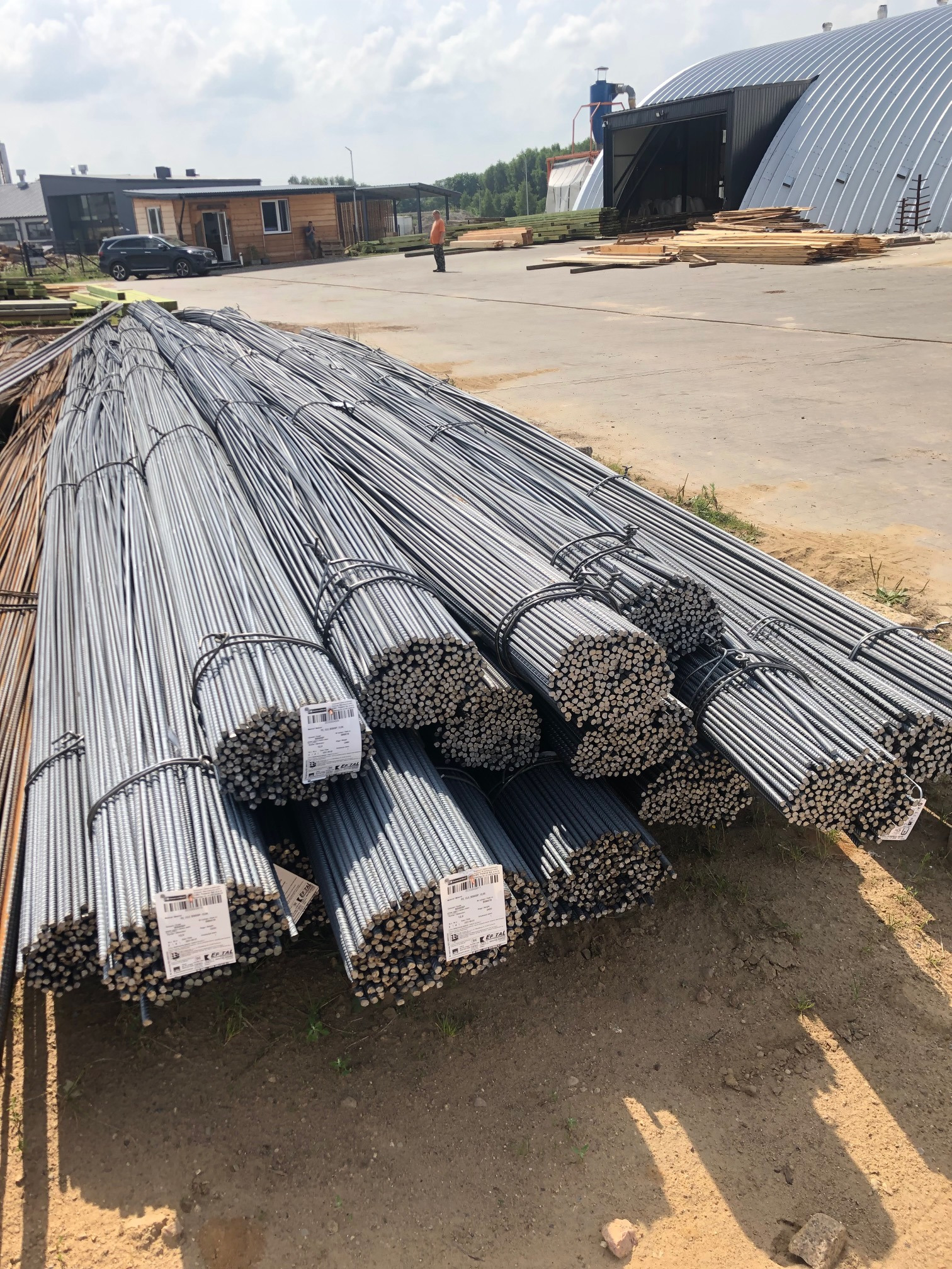 Sztaple wiązek prętów zbrojeniowych z etykietami producenta, ułożone na placu budowy, w tle budynki i osoba w odblaskowej kamizelce.