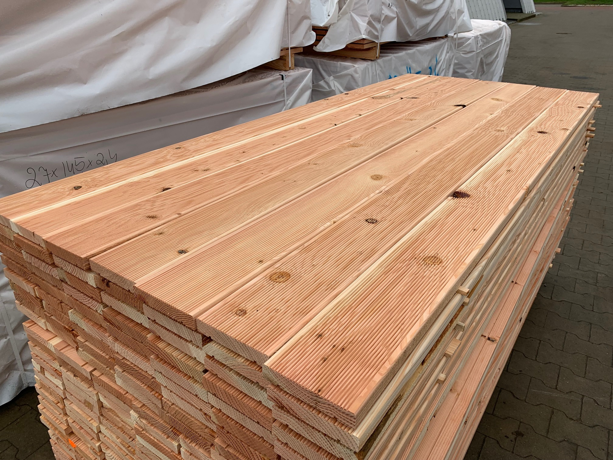 Stos desek tarasowych z jasnego drewna, widoczne słoje i sęki, pakowane w białą folię ochronną, ułożone na zewnątrz.