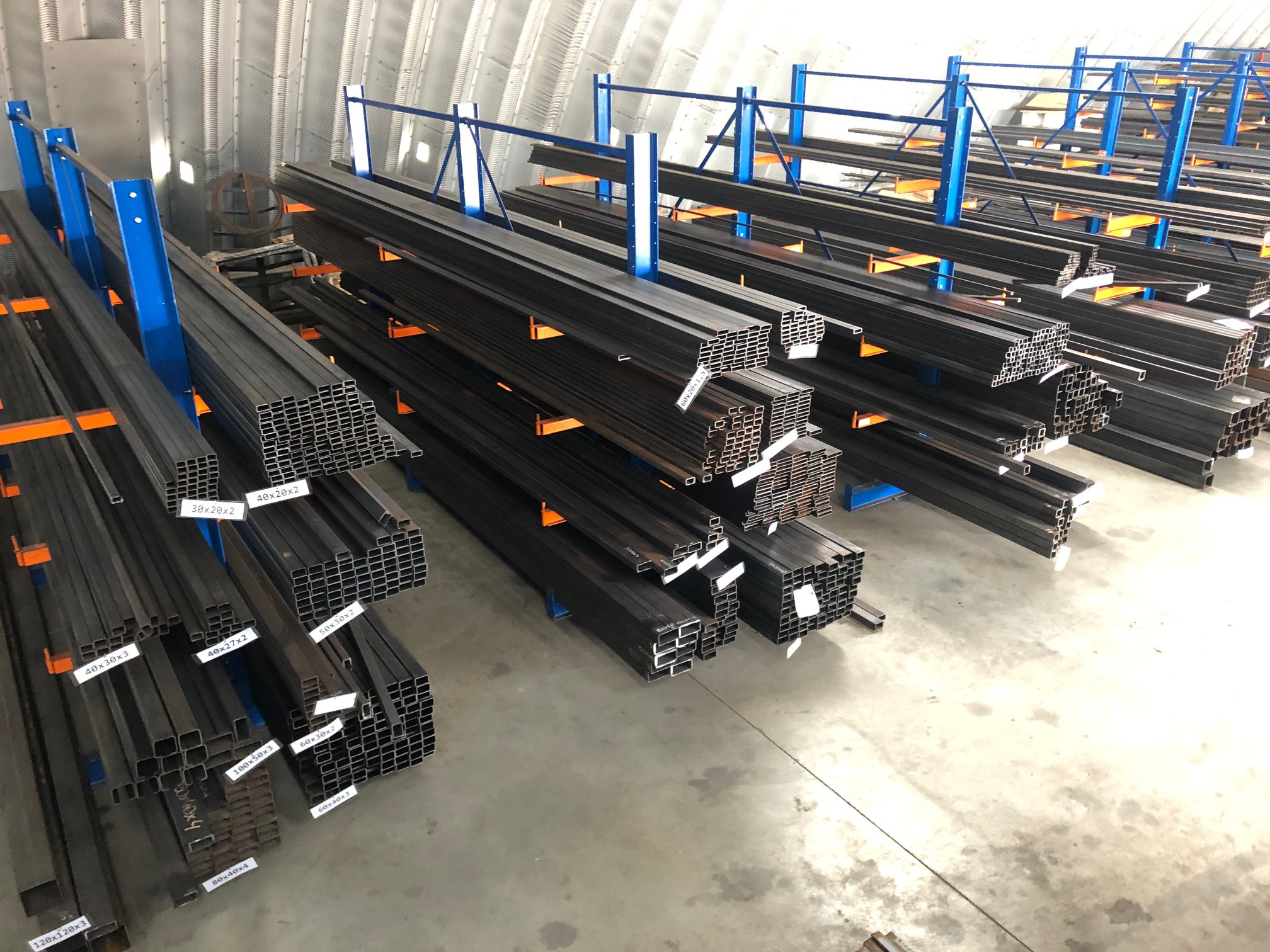 Magazyn profili stalowych kwadratowych, ułożonych na regałach z niebieskimi wspornikami i pomarańczowymi poprzeczkami, z etykietami wskazującymi wymiary.