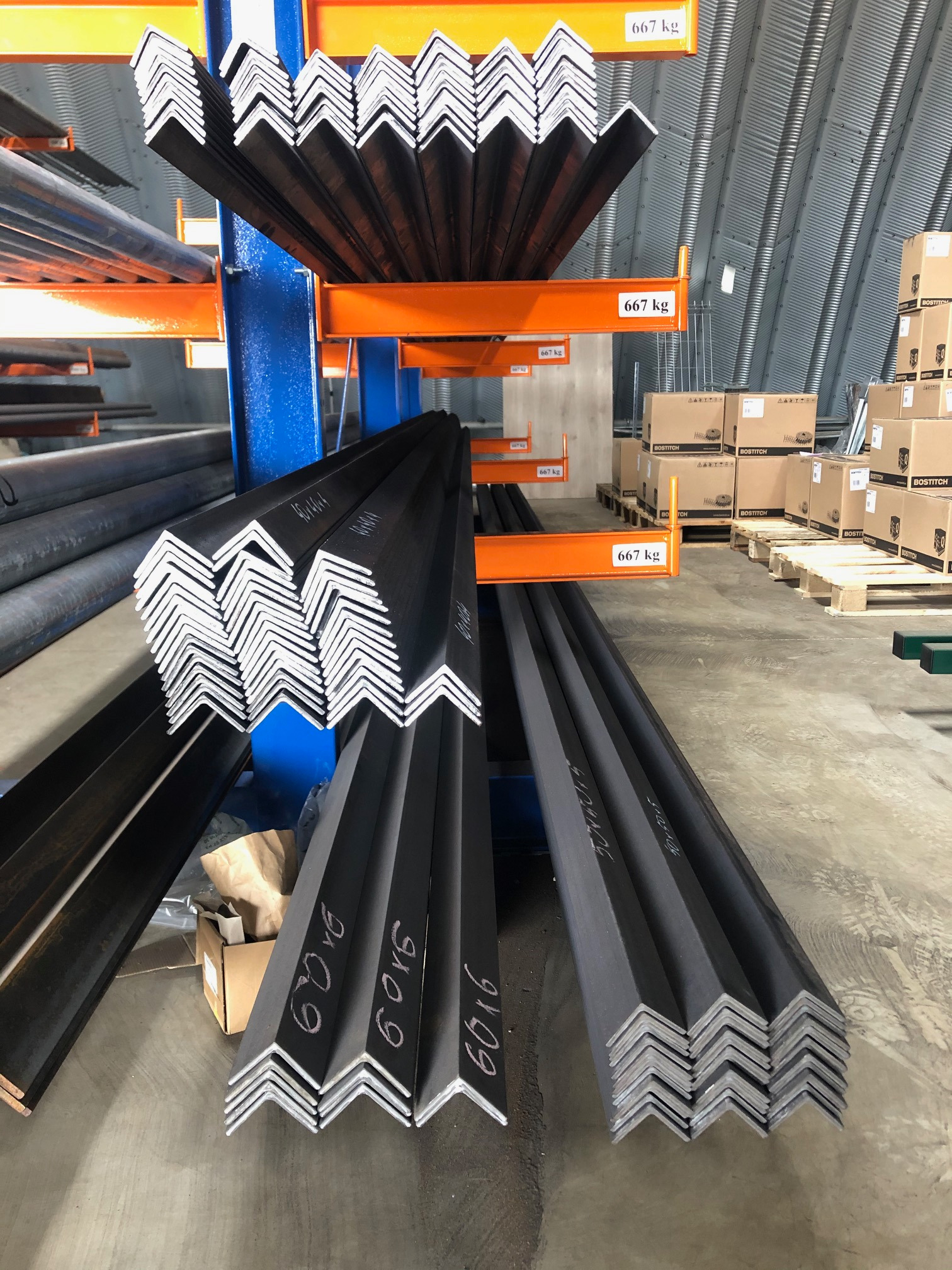 Stalowe kątowniki z białymi oznaczeniami na regale magazynowym, widoczne palety z kartonami w tle.