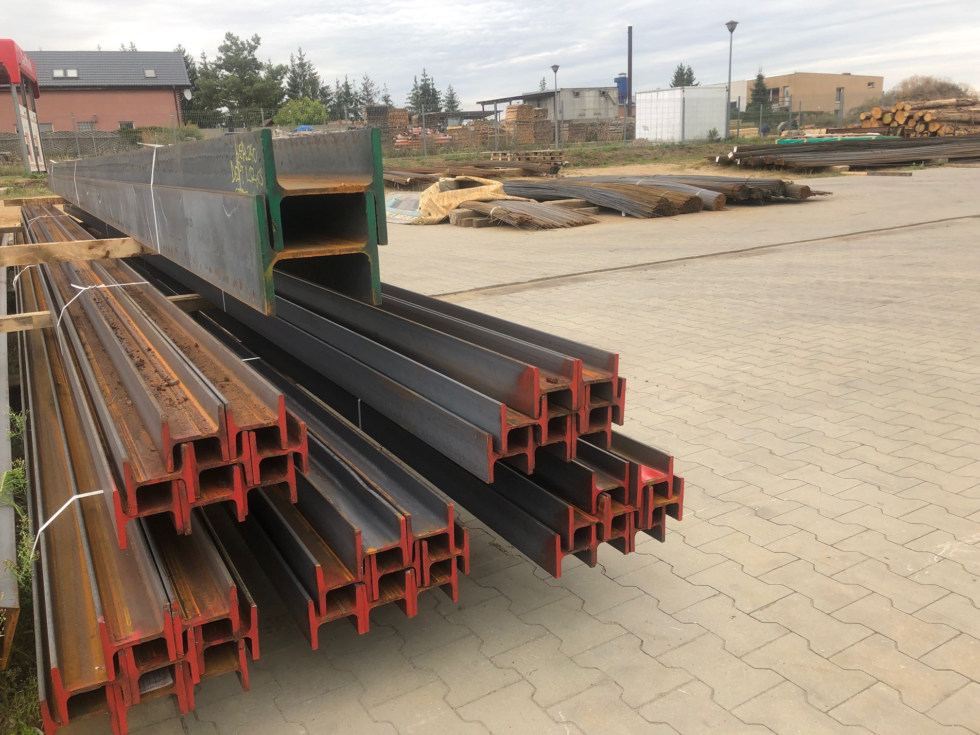 Stalowe belki H-kształtne, pomalowane na czerwono na końcach, ułożone w stos na placu budowy, w tle składowane drewno i budynki.