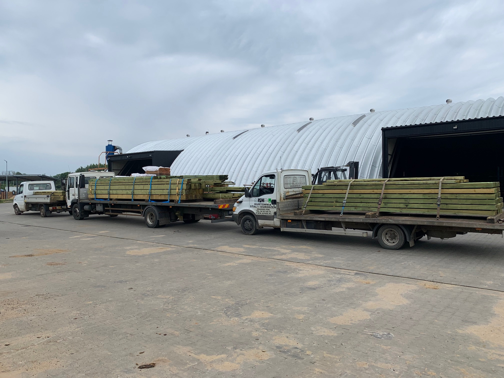 Ciężarówki z przyczepami załadowane drewnem, na tle magazynu z charakterystycznym półokrągłym dachem.