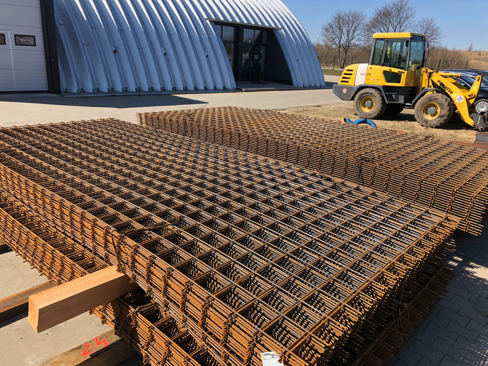 Stosy siatek zbrojeniowych na drewnianych legarach, w tle hala magazynowa o łukowej konstrukcji i żółta ładowarka Komatsu WA75.