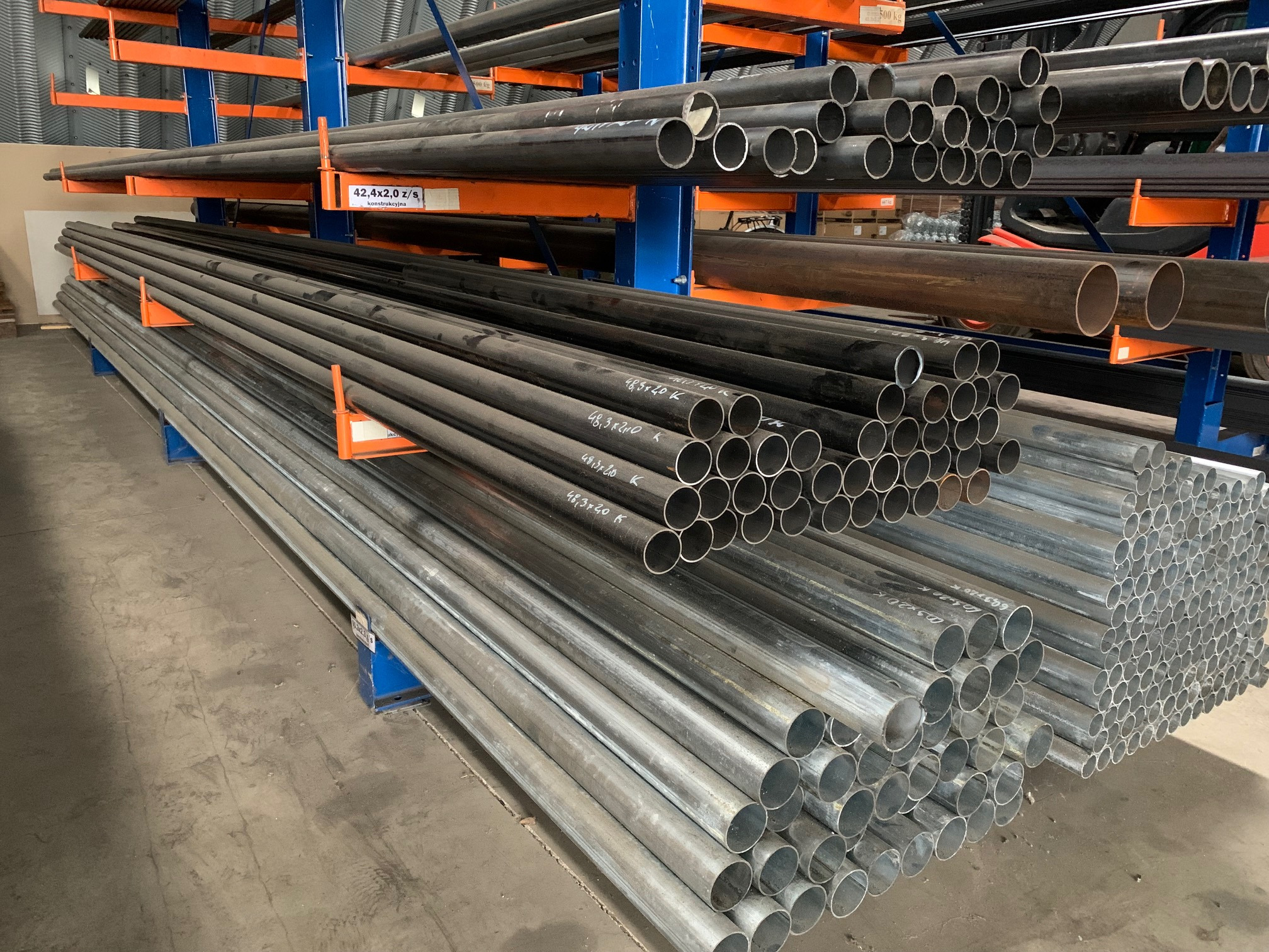 Stalowe rury o różnych średnicach, ułożone na metalowym regale w magazynie. Widoczne oznaczenia na rurach, takie jak wymiary i inne informacje techniczne. Rury w kolorach szarym i czarnym.