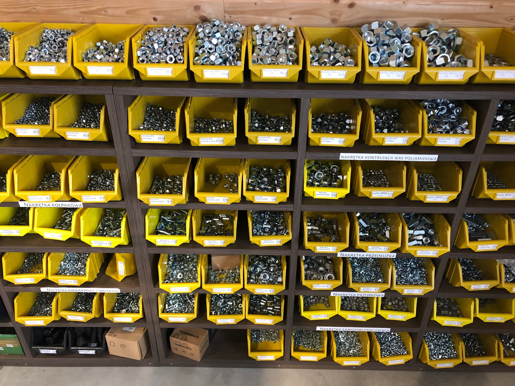 Półki regału w sklepie z metalowymi nakrętkami, śrubami i podkładkami, posegregowanymi w żółtych pojemnikach z etykietami opisującymi rodzaj i cenę.
