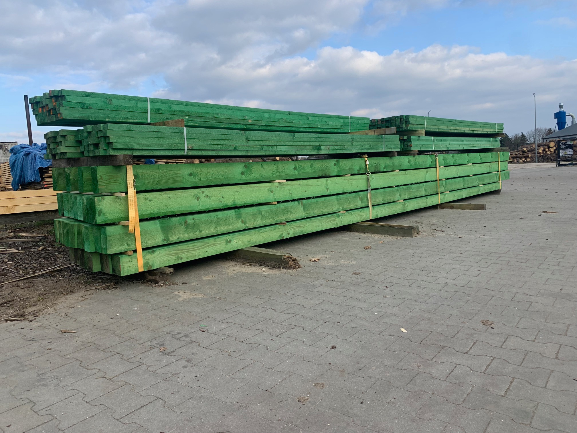 Stos impregnowanych na zielono belek drewnianych, zabezpieczonych żółtą taśmą, składowanych na placu firmy w Drawsku Pomorskim, widok z boku, w tle błękitne niebo z chmurami.