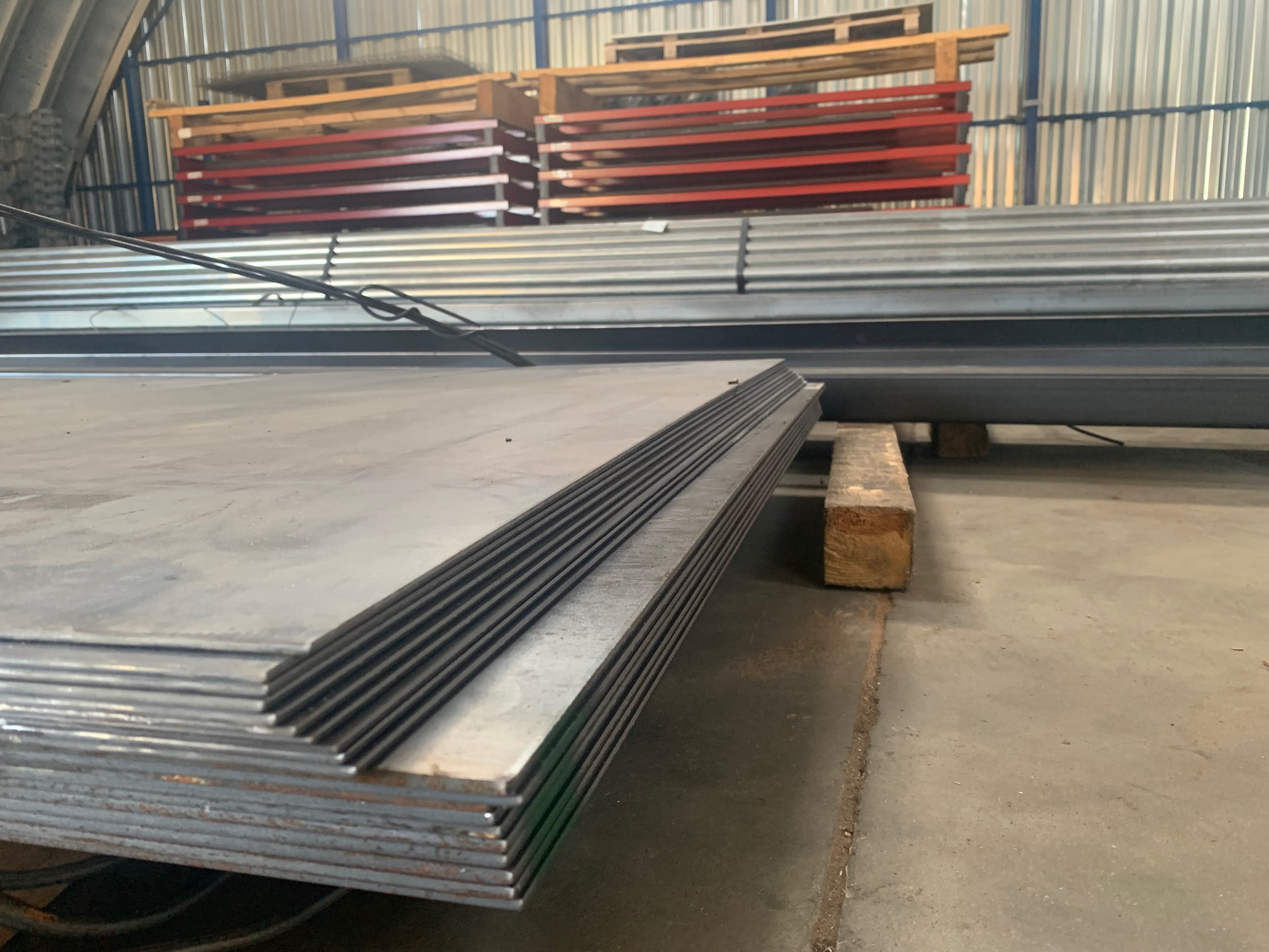 Stos arkuszy blachy stalowej na drewnianej podkładce w magazynie, w tle ułożone palety i profile stalowe.