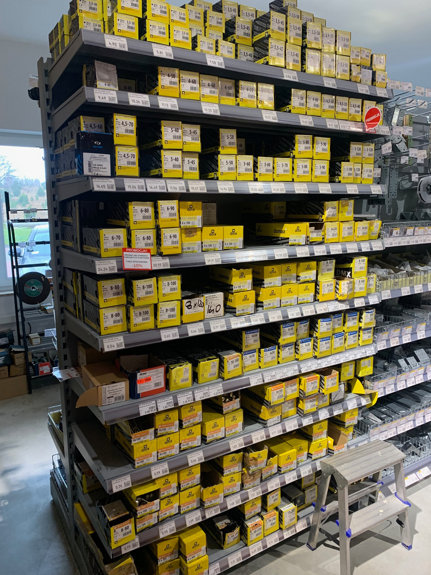 Półki w sklepie budowlanym wypełnione żółtymi pudełkami z łącznikami do drewna, widoczne ceny i oznaczenia producenta DMX, w tle drabinka.