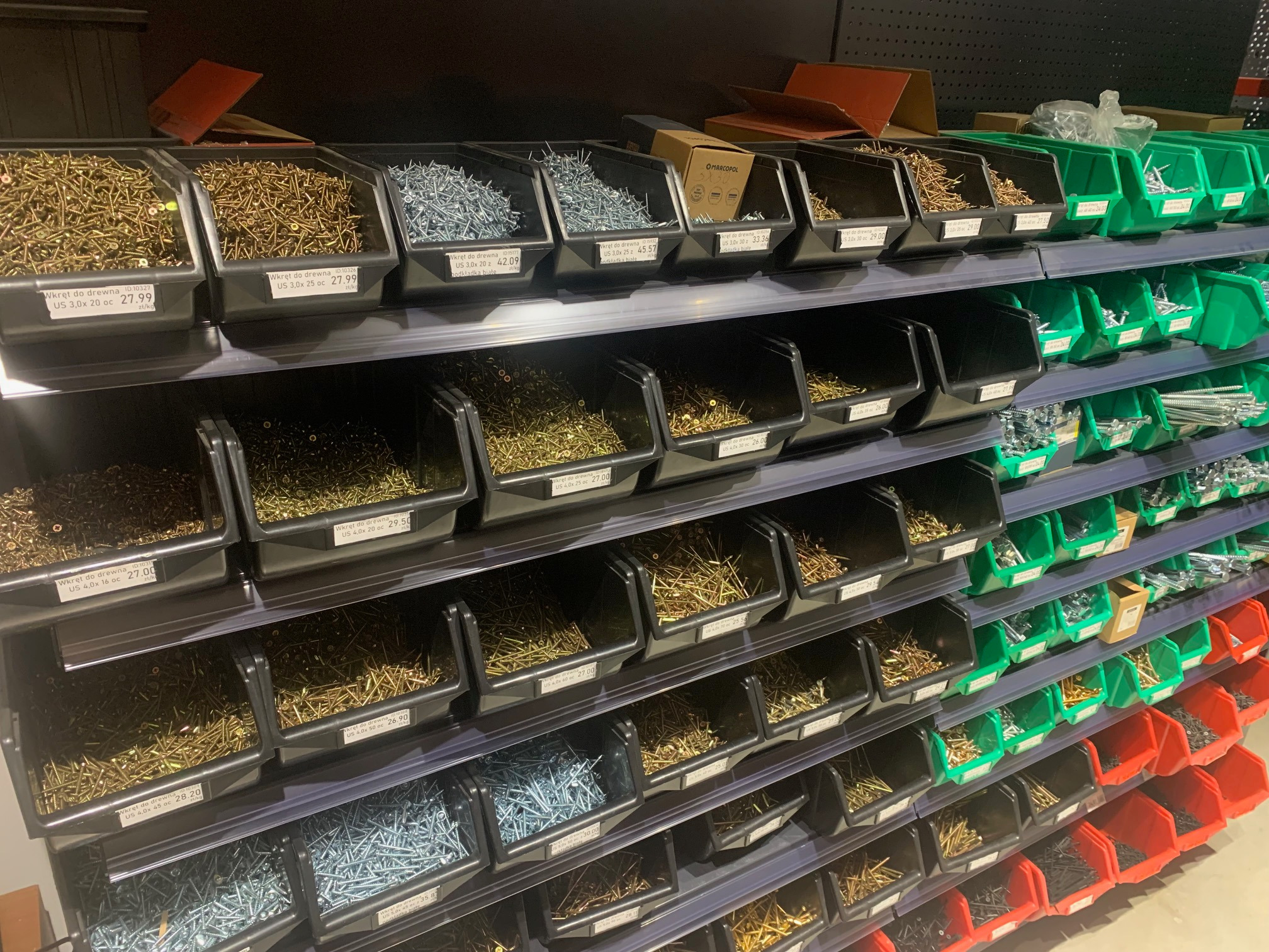 Półki w sklepie budowlanym z pojemnikami pełnymi różnych rodzajów wkrętów do drewna, pogrupowanych kolorami pojemników: czarne, zielone, czerwone, z etykietami cenowymi i opisami.