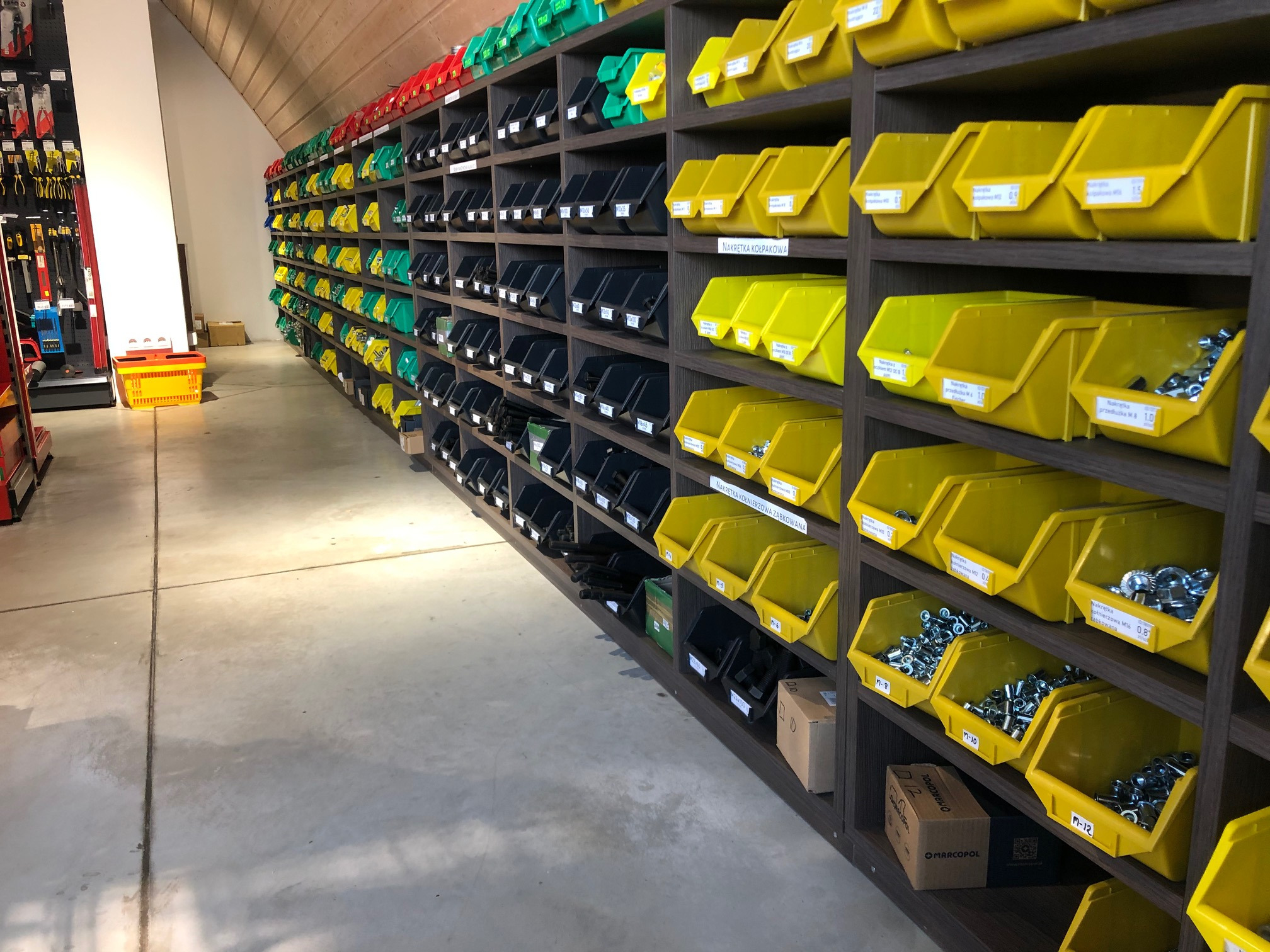 Półki regału w sklepie z artykułami metalowymi, wypełnione kolorowymi pojemnikami z nakrętkami, śrubami i podkładkami, z etykietami informacyjnymi na każdym pojemniku.