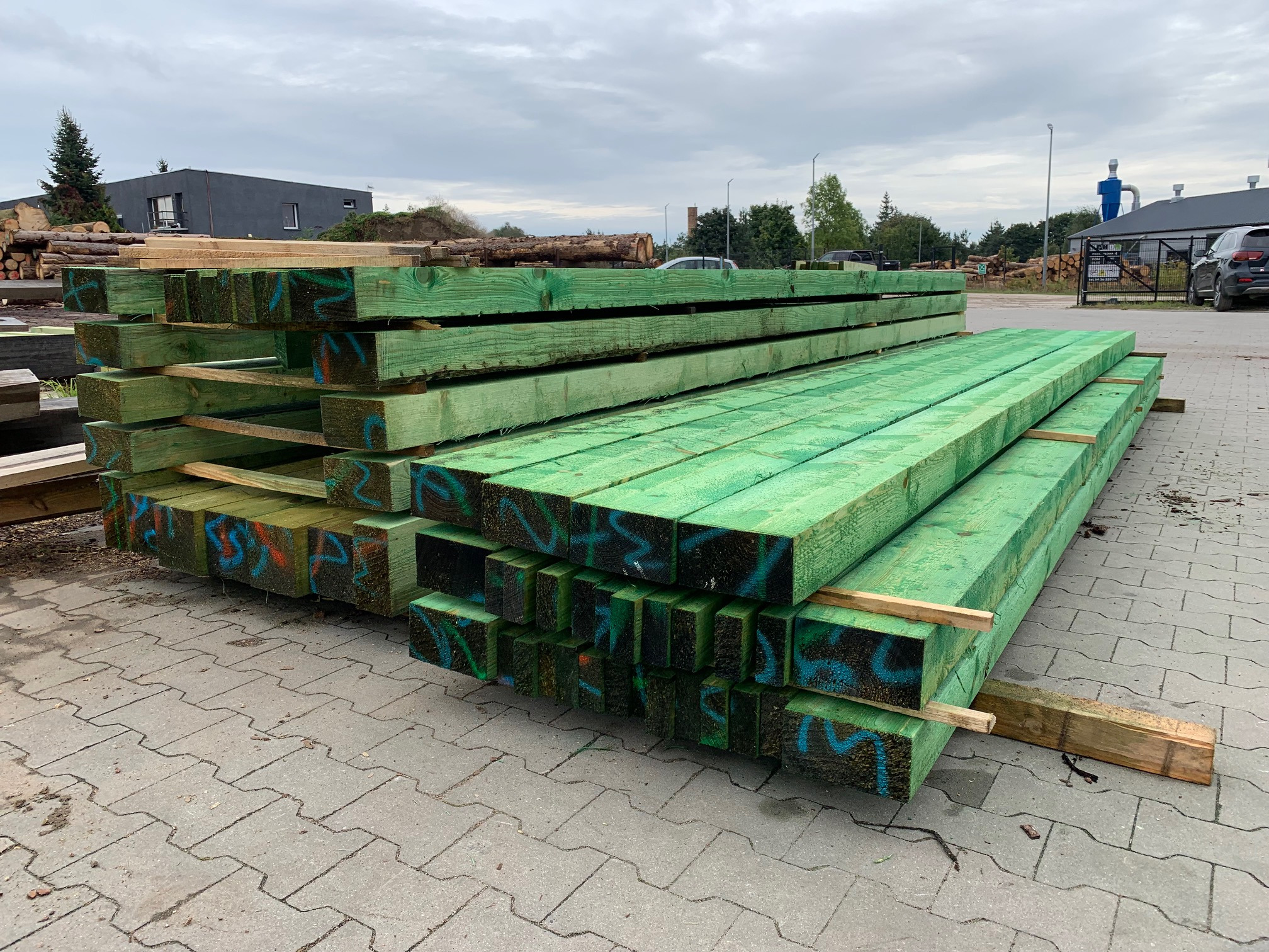 Stosy impregnowanego na zielono drewna konstrukcyjnego na placu, z widocznymi oznaczeniami w kolorze niebieskim i pomarańczowym na końcach.