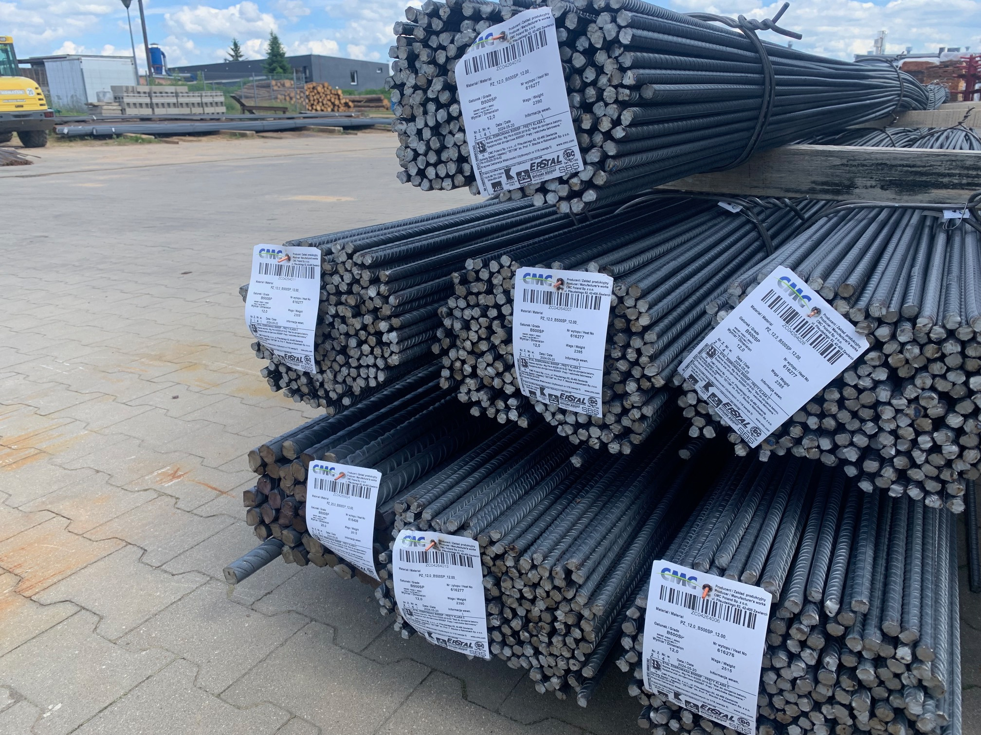 Stosy żebrowanych prętów stalowych ułożone na palecie, widoczne etykiety producenta z informacjami o produkcie, tło stanowi plac budowy z elementami ciężkiego sprzętu.