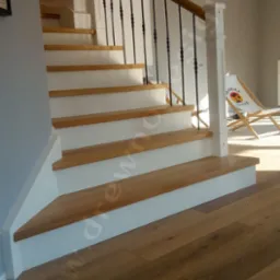 Drewniane schody z białymi podstopnicami i czarną, kutą balustradą, widoczne z dołu, z jasną podłogą i leżakiem 'Bahama House' w tle.