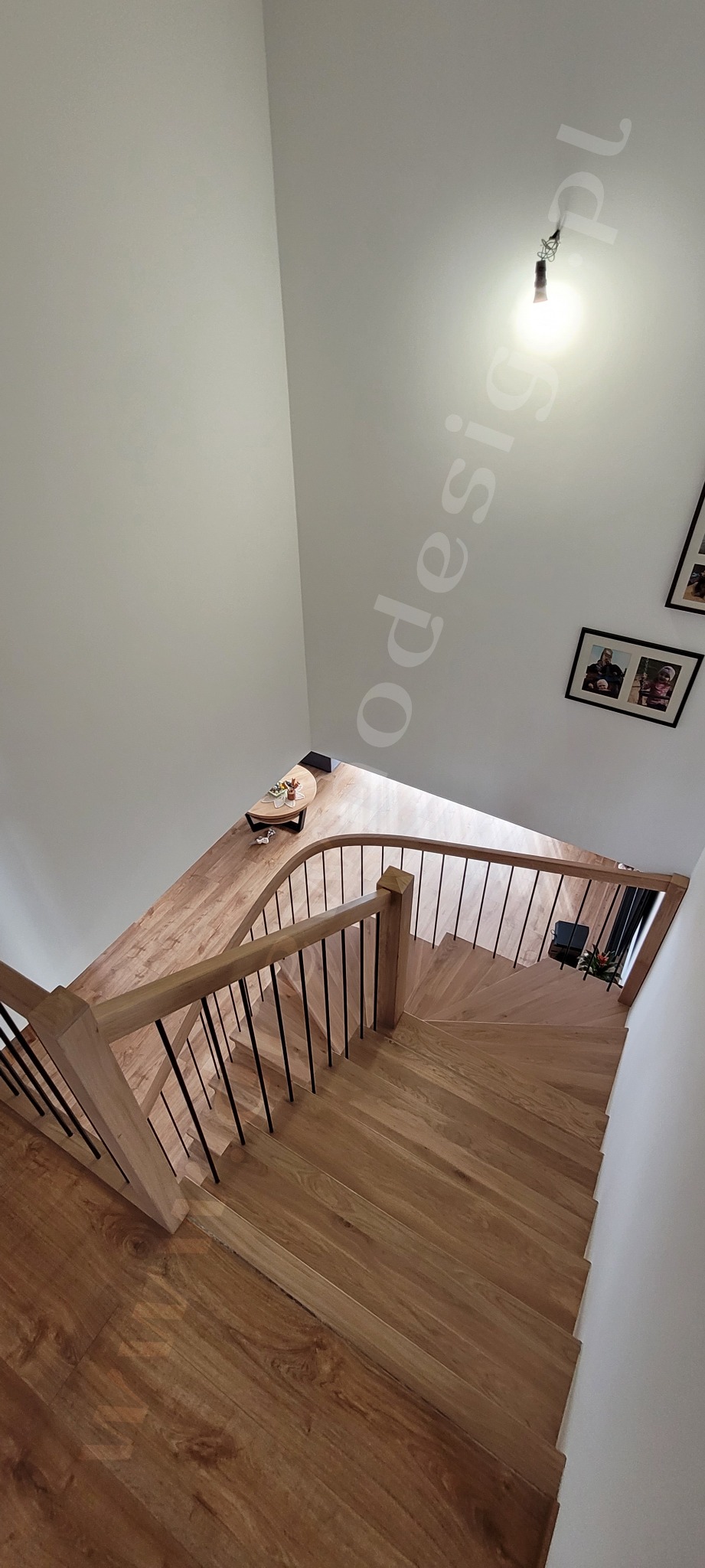 Kątowe ujęcie drewnianych schodów z czarnymi metalowymi prętami balustrady, widok z góry na drewniane stopnie i podłogę, w tle stolik kawowy i zdjęcia na ścianie.