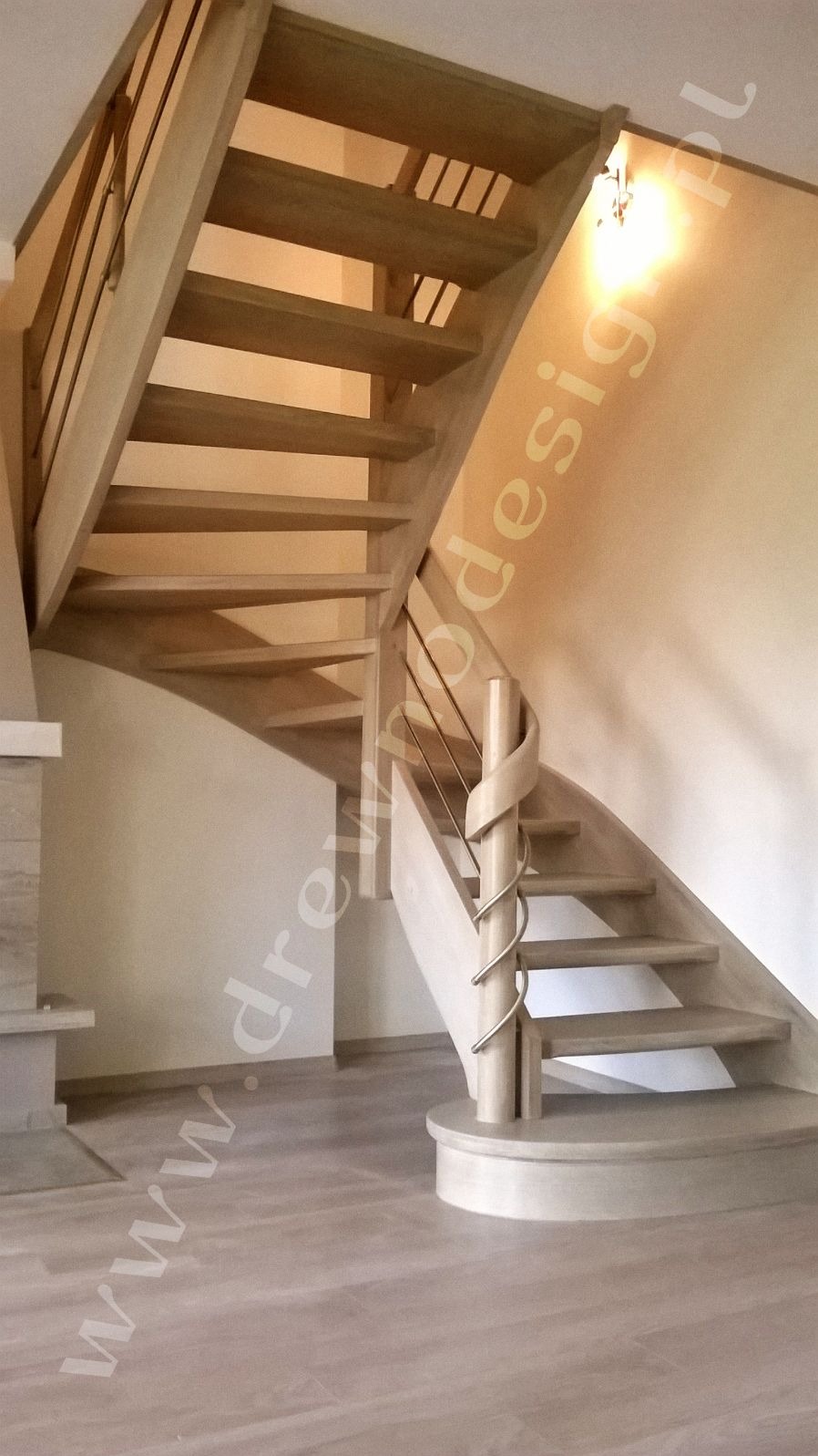 Jasne, drewniane schody z metalowymi poręczami, zakręcające w prawo, z dekoracyjnym słupkiem na dole, widok z dołu.